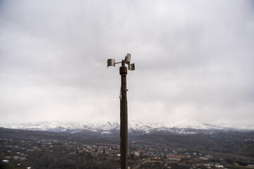 A solitary wind sensor perched atop a slender pole, stark against an overcast sky, silently monitors atmospheric conditions above an industrial complex. Its simplicity belies its importance in data co