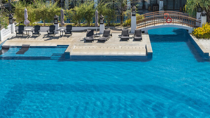 Top view of a large swimming pool with clear blue water. An arched pedestrian bridge spans to a terrace for relaxing with armchairs, sun loungers, tables. A lifebuoy on the railing. Green vegetation