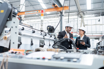 An American male engineer and an Asian female executive inspect the production of materials from machines in an automobile factory. Wear a safety helmet and suit. Use a laptop for work