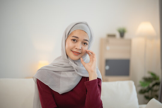 Muslim Islam woman wearing hijab or grey scarf sits on a couch. She is smiling and looking at the camera