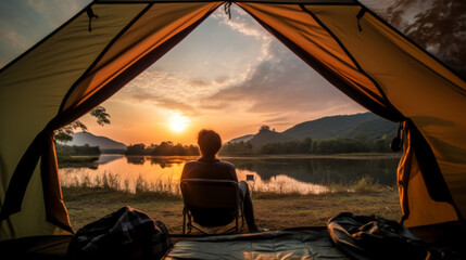 A man sits in a chair near a tent and looks at the water. The sun sets, flooding the area with warm light. Traveler enjoys the tranquility of the moment.