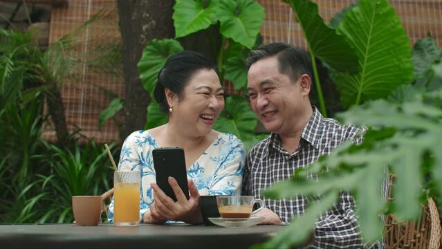 Medium shot of cheerful mature Asian spouses looking at photos of their grandchildren on smartphone sitting at table in outdoor cafe