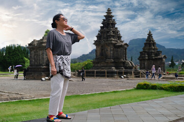 Asian middle-aged tourist standing near Candi Arjuna, an old Buddhist temple at Dieng plateau,...