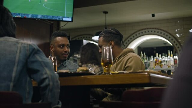 African American men talk sitting in stylish bar together in the evening. Male friends, sport fans having dinner, resting in sports bar on weekend. TV screen with live football match in the background
