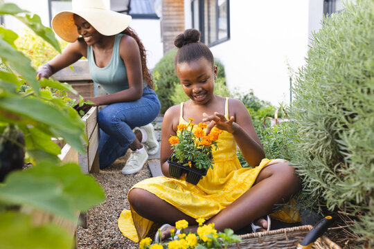 A young African American girl admires a potted flower in the garden at home, with her mother behind 