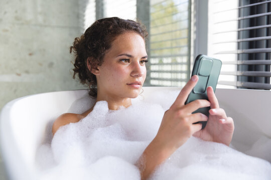 A young biracial woman relaxes in a bubble bath at home, using her smartphone