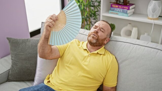 Middle-aged caucasian man suffering in hot weather, fanning himself with handfan while sitting on sofa at home