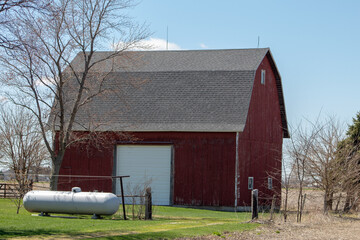 A large propane tank in front of a barn on farm land.