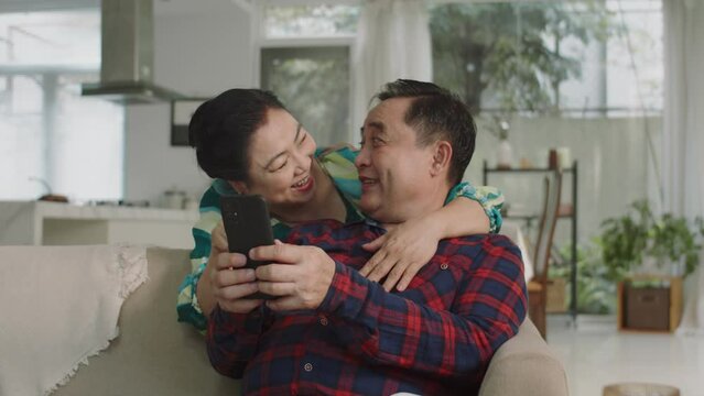 Medium shot of cheerful Asian aged spouses cuddling each other while learning to take selfie on smartphone at home