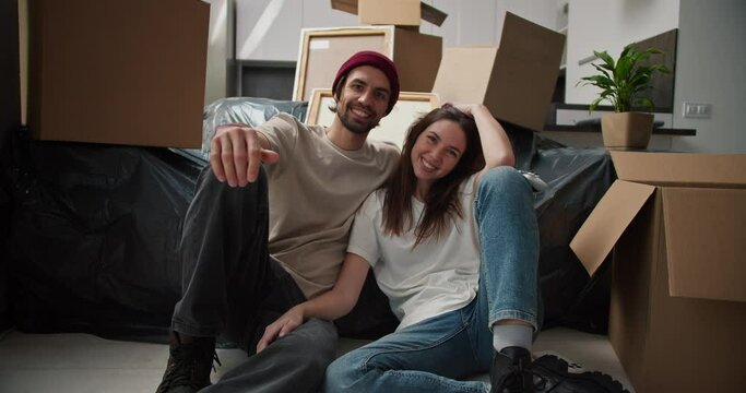 Portrait of a happy brunette man with stubble in a red hat and a beige T-shirt who sits on the floor with his brunette girlfriend in a white T-shirt near the sofa in a black cellophane case among a