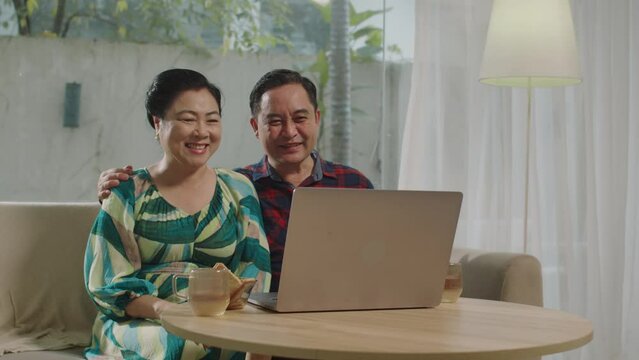 Medium long shot of cheerful senior spouses sitting in front of laptop while talking on video with grandkids