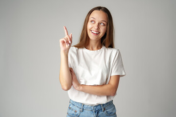 Portrait of pretty young woman in white T-shirt pointing up on gray background