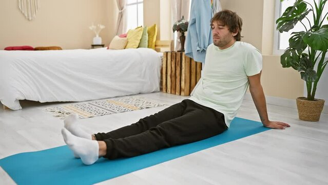 A relaxed young man sits on a yoga mat in a bright home bedroom setting, surrounded by cozy decor.