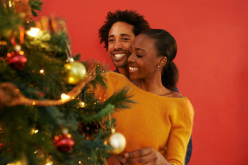 Couple, Christmas and tree decoration in home with happiness for vacation celebration, giving or bonding. Man, woman and smile with lighting for winter season for family event, embrace or lounge