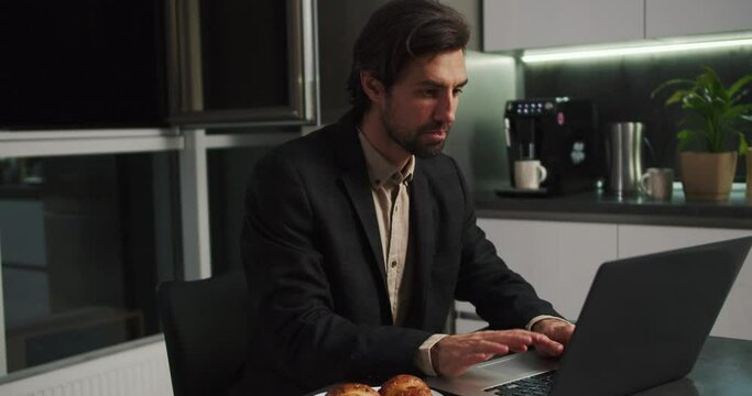 A serious brunette man with stubble in a black suit sits at a table in front of a laptop and communicates via video conference while sitting without pants only in shorts in a modern kitchen in the