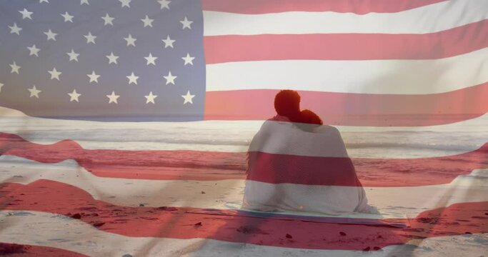 Animation of american flag over happy diverse couple wrapped in rug embracing sat on sunset beach