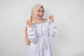 Overjoyed young Asian Muslim woman wearing hijab and white dress clenching fist winner gesture, isolated on white background. Ramadan and Eid Mubarak concept