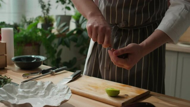 Cropped shot of unrecognizable male chef in striped apron peeling fresh ripe green avocado with spoon, standing by wooden cooking table in kitchen
