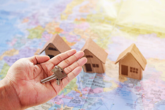  Human hand holding key of paper house toy or cardboard house on world map with morning sunlight.
