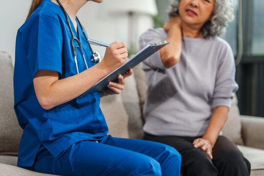 Caucasian teenager female doctor consults with and Asian elderly patient on medical history, discussing headaches, chest pain, coughing, sneezing, stomach pain, and neck pain while seated on a sofa.