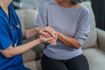 Caucasian female doctor offers encouragement to Asian mature elderly patient woman while holding hands, providing comfort and support during a medical consultation at sofa.