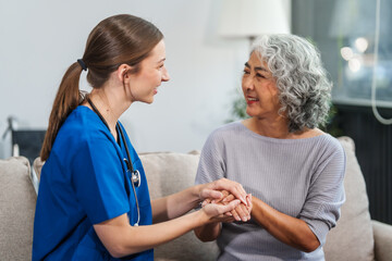 Caucasian female doctor offers encouragement to Asian mature elderly patient woman while holding hands, providing comfort and support during a medical consultation at sofa.