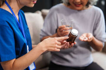 Caucasian female doctor provides a glass of water and medication pills to an elderly Asian patient while they are seated together on the sofa.