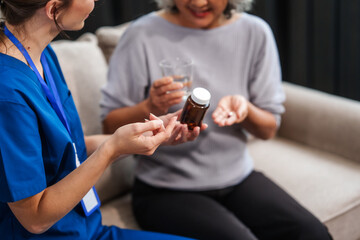 Caucasian female doctor provides a glass of water and medication pills to an elderly Asian patient while they are seated together on the sofa.