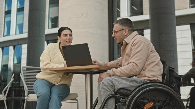 Young businessman in wheelchair discussing project on laptop to female colleague while having meeting at outdoor coworking space in city downtown