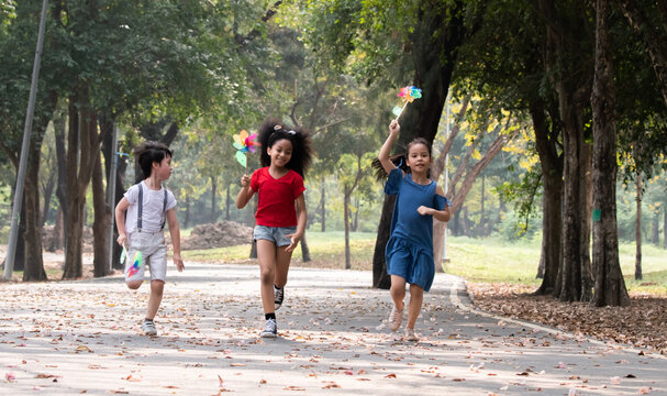 Joyful multiethnic children sprinting in a park, with windmills in hands, laughing happiness on sunny day, young school kids' friends enjoy outdoor activities running and playing together on weekend