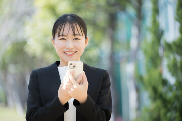 新緑の中、外でスマホを見る若いスーツの日本人(アジア人)女性　カメラ目線