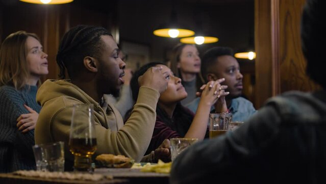 Supportive fans getting upset and angry when soccer team loses world league tournament. Group of multiethnic friends watch football match broadcast in bar or sports pub in the evening, discuss game.