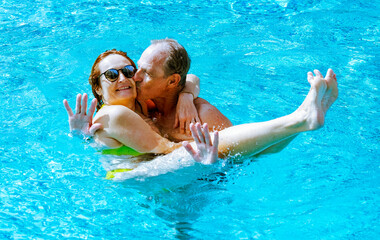 Bonding lovely mature caucasian couple having fun floating in outdoor swimming pool under the sun. Vacation, healthy lifestyle and relax concept