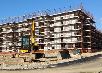 Front view of compact excavator at work on the dirt area with a large builing under construction on behind.