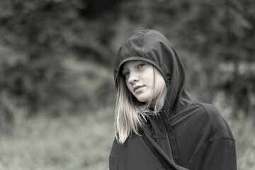 Portr&auml;t junges h&uuml;bsches M&auml;dchen in Regenjacke mit Kapuze. Bei grauem wolkenverhangenen Himmel geht das M&auml;dchen durch den Wald spazieren. Sie hat lange blonde Haare, misstrauisch blickende Augen.