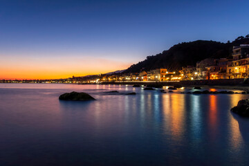 Obraz premium evening or night landscape of evening town coastline in golden lights and sea gulf with calm water and nice reflections with beautisul sunset sky on background