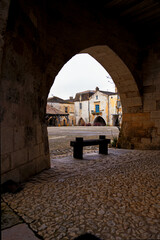 Portrait of the Place des Cornières in Monpazier with an arch and wooden bench in the foreground...