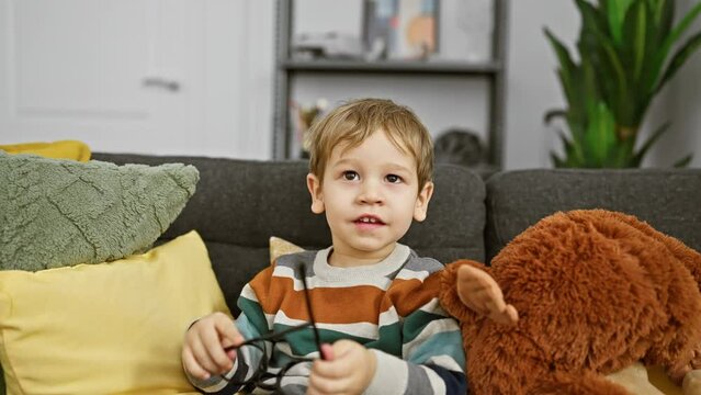 Blond toddler boy plays with glasses on a couch, with a teddy bear, in a cozy living room.