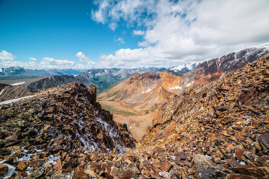 Top view from abyss edge between rocks to multicolor valley of iron colors and big sharp rocky ridge of red color. Colorful large mountains in freshly fallen snow in low clouds. Vivid alpine scenery.