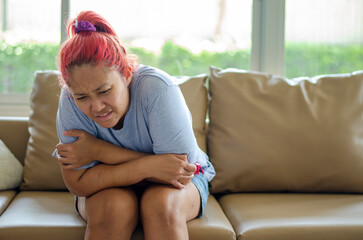 Young Asian woman has menstrual cramps or stomach pain from other causes. She sits on the sofa.