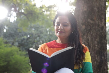 Portrait of mid aged woman holding a black color almanac in hand, reading personal almanac. sunlight, green park environment. book promotion, blank cover. - obrazy, fototapety, plakaty