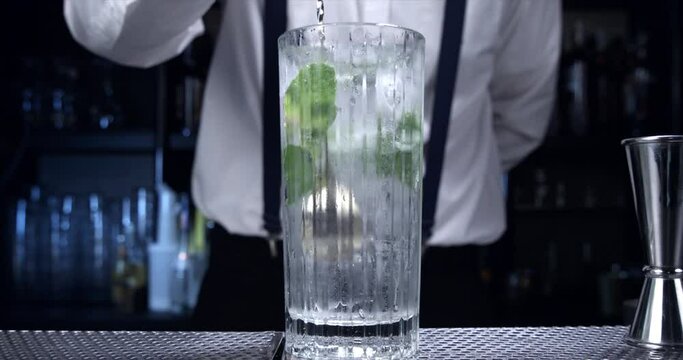 Super slow motion macro of bartender is mixing fresh aromatic leaf of peppermint herb in transparent glass with soda and ice cubes during mojito cocktail preparation in bar or restaurant at 1000 fps.