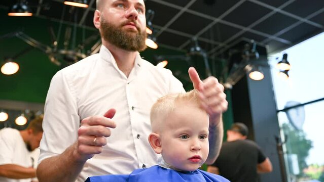 Young child getting haircut in modern barbershop. Hairdresser making final touches in new haircut, shaping child's hair by hands.