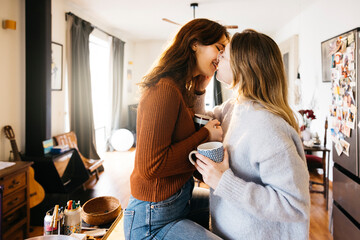 Young female couple kissing while sharing a cup of tea in a in their home kitchen. Lesbian couple...