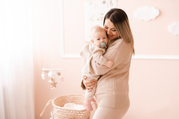 Happy mother holding newborn baby boy with pacifier in room over beige. Motherhood.