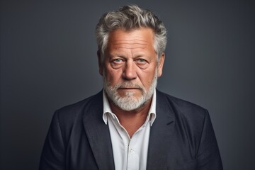 Portrait of a serious senior man with grey hair and beard standing against grey background.