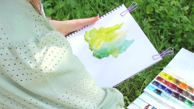 Female artist painting still life on plein air in park,. Woman drawing watercolor sketch and sitting on grass outdoor. Girl in green summer dress sketching, top view from the side