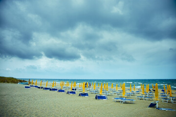 Empty beach chairs and umbrellas await visitors on a sandy shore, as a tempestuous sky looms...