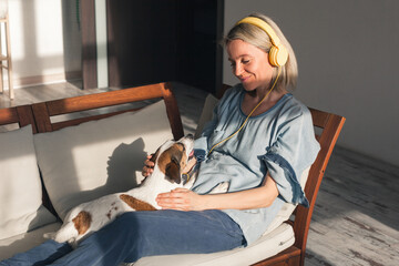Happy middle aged woman wear earphones relax on couch listen to music on smartphone