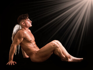 A man without clothes sits on the ground, his angel wings spread out behind him. The man appears relaxed as he gazes into the distance. - 758815119